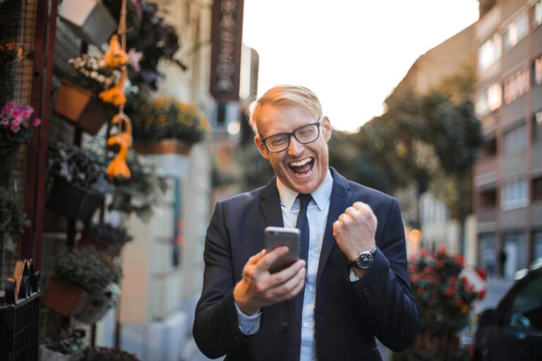 A joyful man in a suit celebrates good news on his smartphone in an urban street setting.